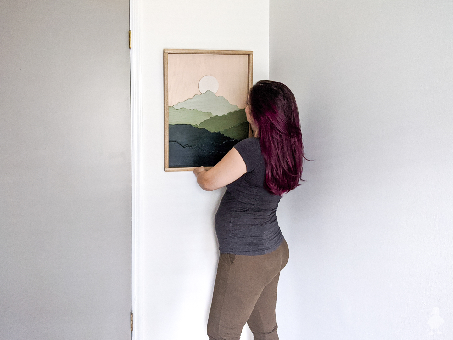 woman hanging up scroll saw art of green mountains in the nursery on a white wall with a wood frame